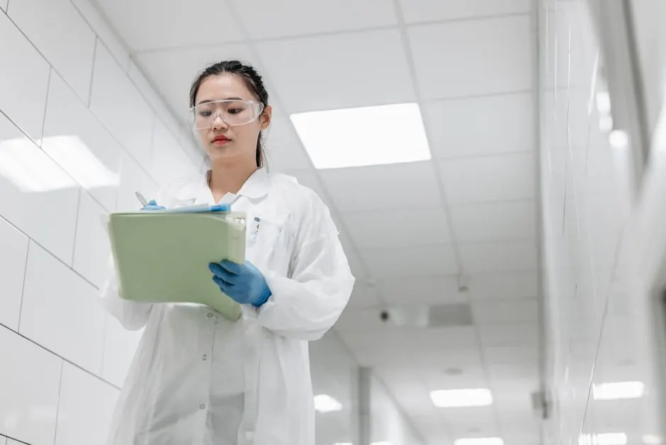A focused female scientist in a lab coat writes notes in a laboratory hallway, showcasing professionalism in research settings.
