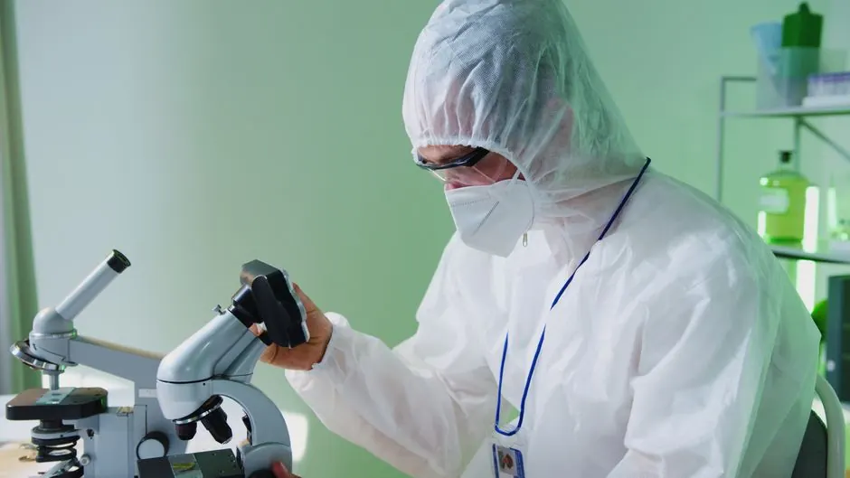 A scientist in protective gear examines samples under a microscope in a lab.